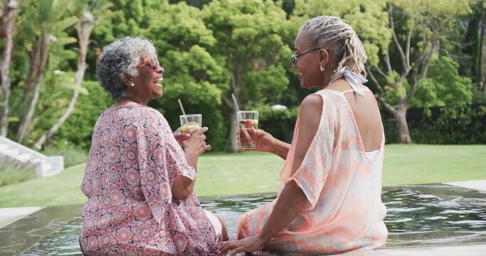 Happy senior african american female friends making a toast with drinks sitting by pool, slow motion
