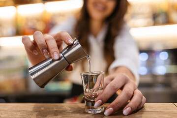 Beautiful female bartender pouring alcohol into shot glass shelves full of bottles with alcohol on the background