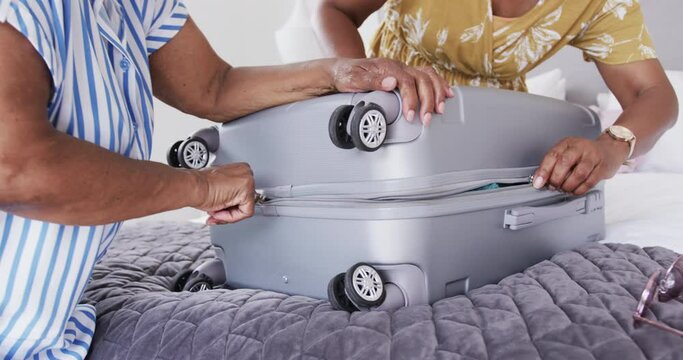 Senior african american female friends packing suitcase on bed at holiday home, slow motion