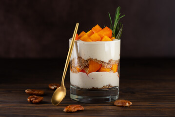 Vegan parfait in a glass with persimmon, rosemary, pecan, whipped cream and spoon on a brown....