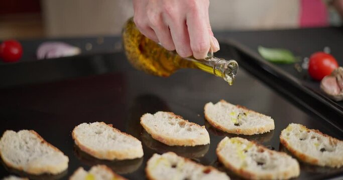 woman pours olive oil onto sliced baguette pieces on a baking tray making croutons or bread chips