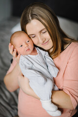 Happy mom embrace infant child enjoying together sitting on a bed at home. A young mother laughing holds a newborn baby son. Tired loving mom on maternity leave. Mother's day concept. Closeup.