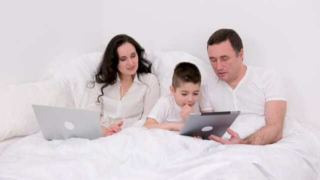 husband wife in bed with little son looking at tablet cartoon interesting game pictures laughing talking communication real people at home love life happy family life joyful people white background