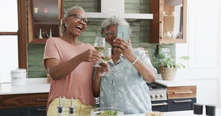 Happy senior african american female friends drinking wine taking selfies in kitchen, slow motion