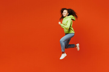 Fototapeta na wymiar Full body side profile view young excited overjoyed woman of African American ethnicity wear green hoody casual clothes jump high run fast isolated on plain red orange background. Lifestyle concept.