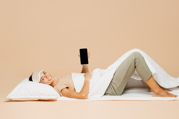 Full body young Latin woman wears pyjamas jam sleep eye mask rest relax at home under duvet use blank screen mobile cell phone isolated on plain pastel beige background. Good mood night nap concept.
