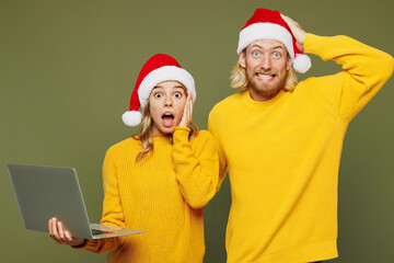 Merry shocked sad young couple two IT friends man woman wear sweater Santa hat posing hold use work on laptop pc computer isolated on plain green background. Happy New Year Christmas holiday concept.