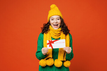 Young surprised happy woman wear green knitted sweater yellow hat scarf hold gift certificate coupon voucher card for store isolated on plain orange red background studio portrait. Lifestyle concept.