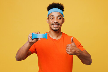 Young happy fitness trainer sporty man sportsman wear orange t-shirt hold energy bar protein snack show thumb up training in home gym isolated on plain yellow background Workout sport fit abs concept