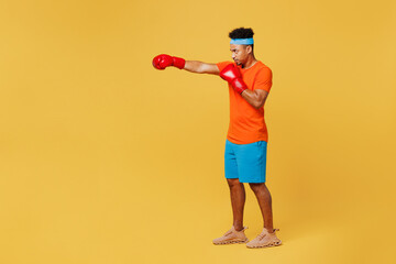 Full body young fitness trainer instructor sporty man sportsman wears orange t-shirt boxing red gloves punch spend time in home gym isolated on plain yellow background. Workout sport fit abs concept.