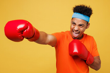 Young masculine fitness trainer instructor sporty man sportsman wear orange t-shirt boxing red gloves punch spend time in home gym isolated on plain yellow background. Workout sport fit abs concept.