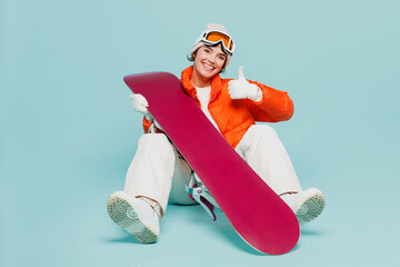 Full body young woman wear warm padded windbreaker jacket hat ski goggles mask sit with snowboard show thumb up travel rest spend weekend winter season in mountains isolated on plain blue background.