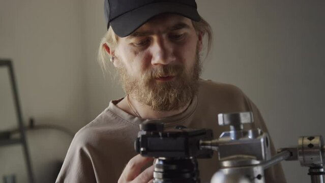 Medium close-up shot of male videographer looking at screen of camera mounted on horizontal rack and adjusting something, while shooting flatlay in studio