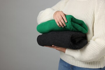 Woman with stack of knitted sweaters on grey background, closeup. Space for text