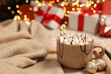Delicious cocoa with marshmallows, wafer sticks and chocolate topping in cup and ornaments against Christmas gifts, closeup. Space for text