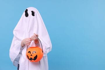 Child in white ghost costume holding pumpkin bucket on light blue background, space for text. Halloween celebration