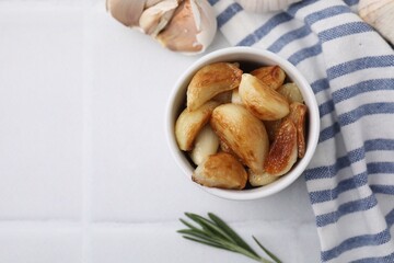 Fried garlic cloves in bowl and rosemary on white table, flat lay. Space for text