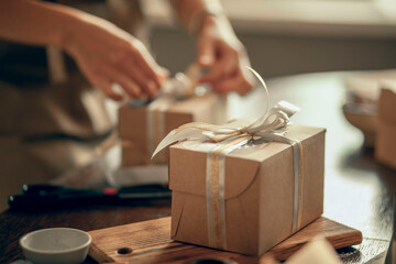 A craft box with white and gold ribbons. A woman's hands ties a ribbon on a customer's order box. A small business entrepreneur and the concept of food delivery. - obrazy, fototapety, plakaty