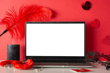 Online intimacy captured in side view scene with a laptop, candle, mask, and adult toys—furry handcuffs and feather tickler on a red background. An empty screen awaits your virtual desires