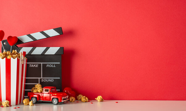 Stay in for a heartwarming movie night on Valentine's Day. Side view picture of a table with a clapperboard, tiny car model, popcorn-filled box, heart decor, and sprinkles against a red wall backdrop