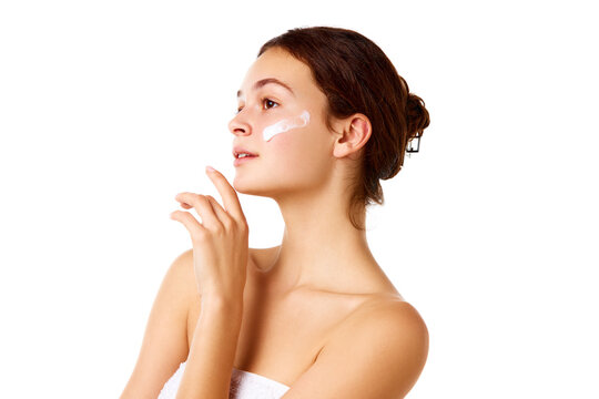 Side view image of young beautiful girl with bare shoulders standing with moisturizing cream on face against white background. Concept of natural female beauty, skin care, cosmetology and cosmetics