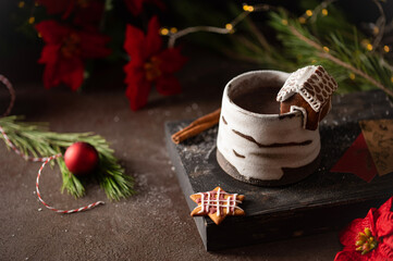 Hot winter drink in a clay mug. Close-up of hot chocolate with cinnamon stick, gingerbread house whipped cream. Winter cozy still life with cocoa, Christmas decorations, fir branch and lights. © Lizaveta