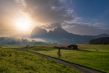 Sunrise over Alpe di Siusi, A morning golden glow bathes the landscape, with the plateau in the background, embraced by fog, and a lush green meadow below