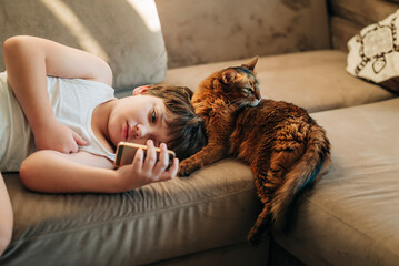 Kid boy watching to his mobile phone lying on the couch sofa with his pet fluffy red Abyssinian cat...