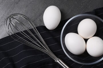 Metal whisk and raw eggs on dark grey table, flat lay