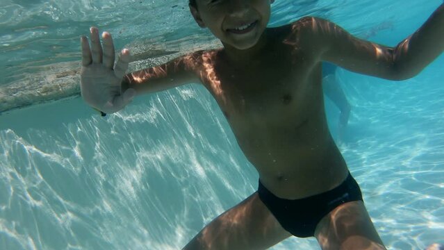 Boy dives underwater, waving hand and smiles in the pool with blue water