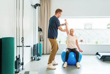 A focused senior woman sits on a blue fitness ball performing arm exercises with the guidance of her physiotherapist in a well-lit rehab gym. - Powered by Adobe