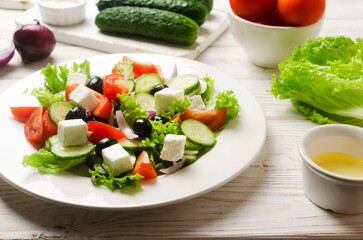 Classic Greek salad with cucumbers, tomatoes, red onion, feta cheese, lettuce and black olives on a white wooden table. The concept of traditional dishes. Healthy food. Simple ingredients.