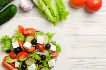 Classic Greek salad with cucumbers, tomatoes, red onion, feta cheese, lettuce and black olives on a white wooden table. The concept of traditional dishes. Simple ingredients. Top view. Copy space