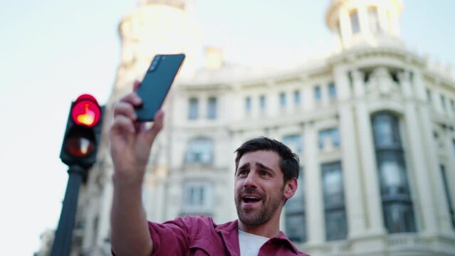 Enthusiastic young man video calling or vlogging in front of historical architecture. His cheerful demeanor and red shirt convey friendly urban travel experience, sharing moment with online audience