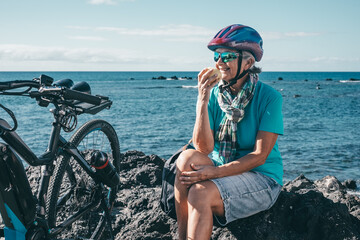 Smiling active senior woman with helmet stops riding bike at sea to eat an apple fruit.  Authentic...