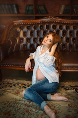 Fototapeta premium Pregnancy. A girl is expecting a baby. Tenderness. New Year. Christmas. Winter. The expectant mother hugs her belly. Beautiful pregnant girl with long red hair. Girl in a white shirt. The girl sits an