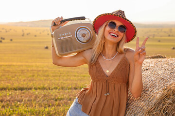 Happy hippie woman with radio receiver showing peace sign near hay bale in field, space for text