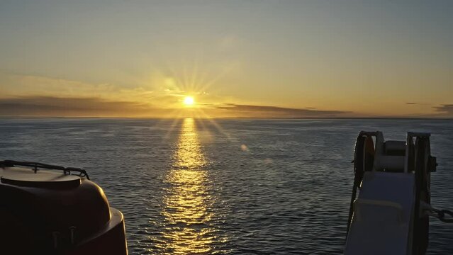 Sunrise at sea starboard deck side view from cruise ship with lifeboat and davit crane. Golden morning sun reflection on the surface with calm waves. On the horizon the coast of Norway, travel concept