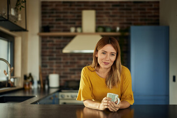 A portrait of a beautiful blonde woman, dressed in a yellow sweater, drinking coffee in the kitchen.