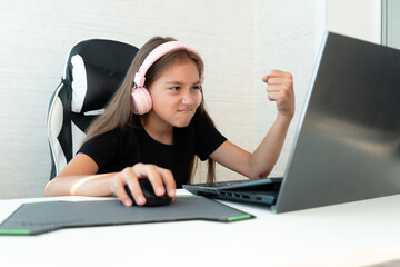 teenage girl in pink headphones is playing computer games on a white background. A negative emotion on the face. the child shows his fist at the laptop screen.