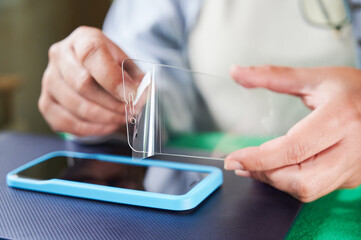 Unrecognizable woman removes a transparent plastic film that covers the anti-scratch screen protector that she is going to install on a smartphone.