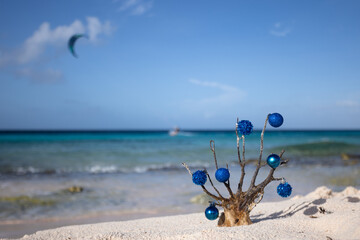 Fototapeta na wymiar Dead corals decorated with blue Christmas balls standing on the sand beach with blue sea behind it, with a kitesurfer away. 