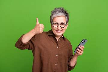 Portrait of satisfied clever person dressed brown clothes in eyewear hold smartphone showing thumb...