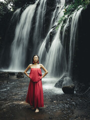 Charming Asian woman posing near waterfall. Nature and environment. Motion water. Travel lifestyle. Beautiful woman wearing long red dress. Slim body. Copy space. Yeh Bulan waterfall, Bali