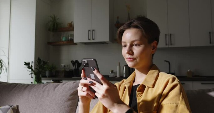 Modern woman using smartphone on sofa, chatting and browsing social media during her free time, searching information on browser wireless internet. Casual lifestyle and comfort. Cinematic advertising