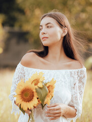 Caucasian woman walking in a field and holding a bouquet of sunflowers. Portrait of smiling woman wearing white dress. Summer vacation. Lifestyle concept. Dreamy mood.