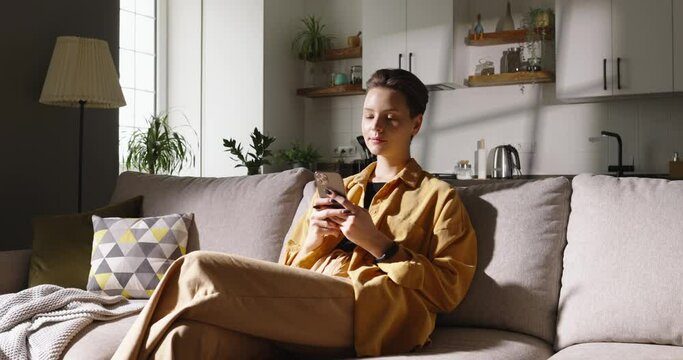 Relaxed caucasian woman in casual clothing sitting on sofa and using smartphone in modern room. Chatting with your boyfriend or friends on phone in free time. Moments of idleness. Cinematic AD