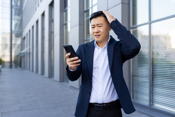 A young Asian man in a business suit is standing outside an office center, looking shocked and...