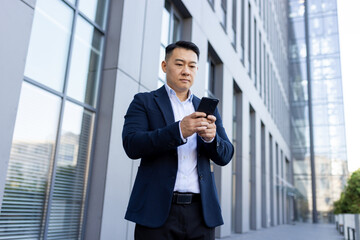 Serious Asian businessman standing on the street near the office with a mobile phone, typing a...