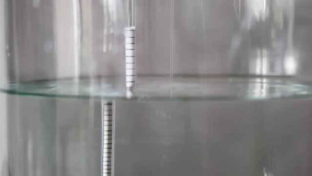 alcohol distilling process closeup - liquid distilled transparent liquid flowing in large glass jar with floating alcoholometer in it.